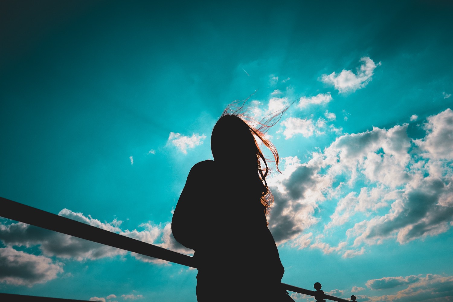 Silhouette of a woman with hair blowing in the wind, standing at a railing against a bright teal sky